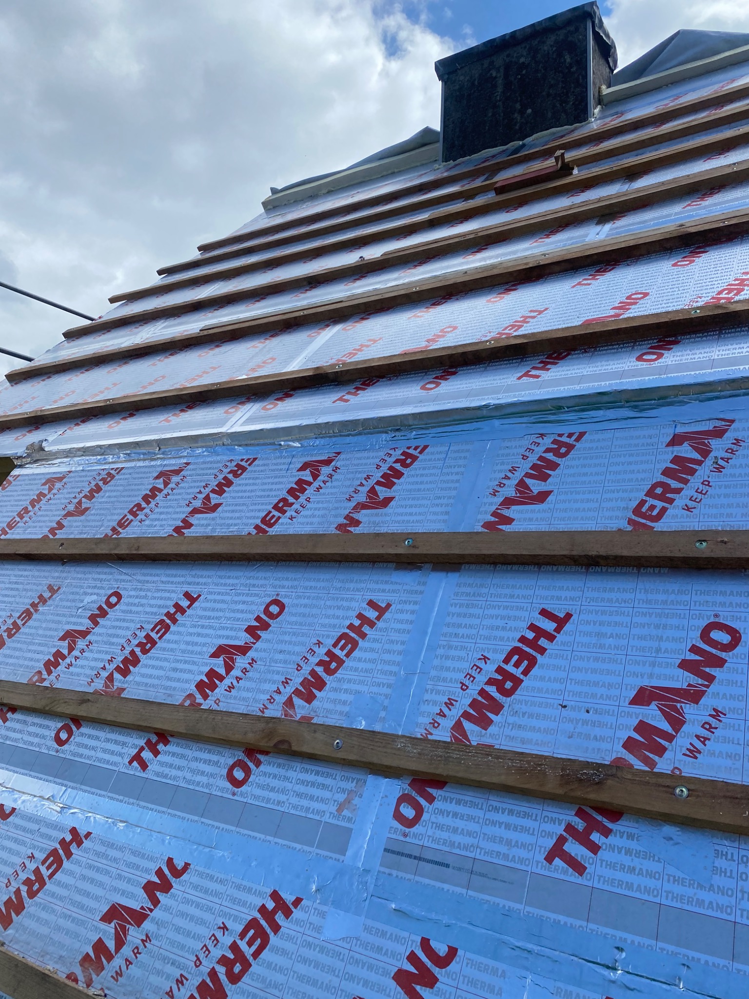 Montaż izolacji termicznej dachu z widocznymi łatami i kontrłatami, folia termoizolacyjna z nadrukiem producenta, widok na komin i pochmurne niebo.