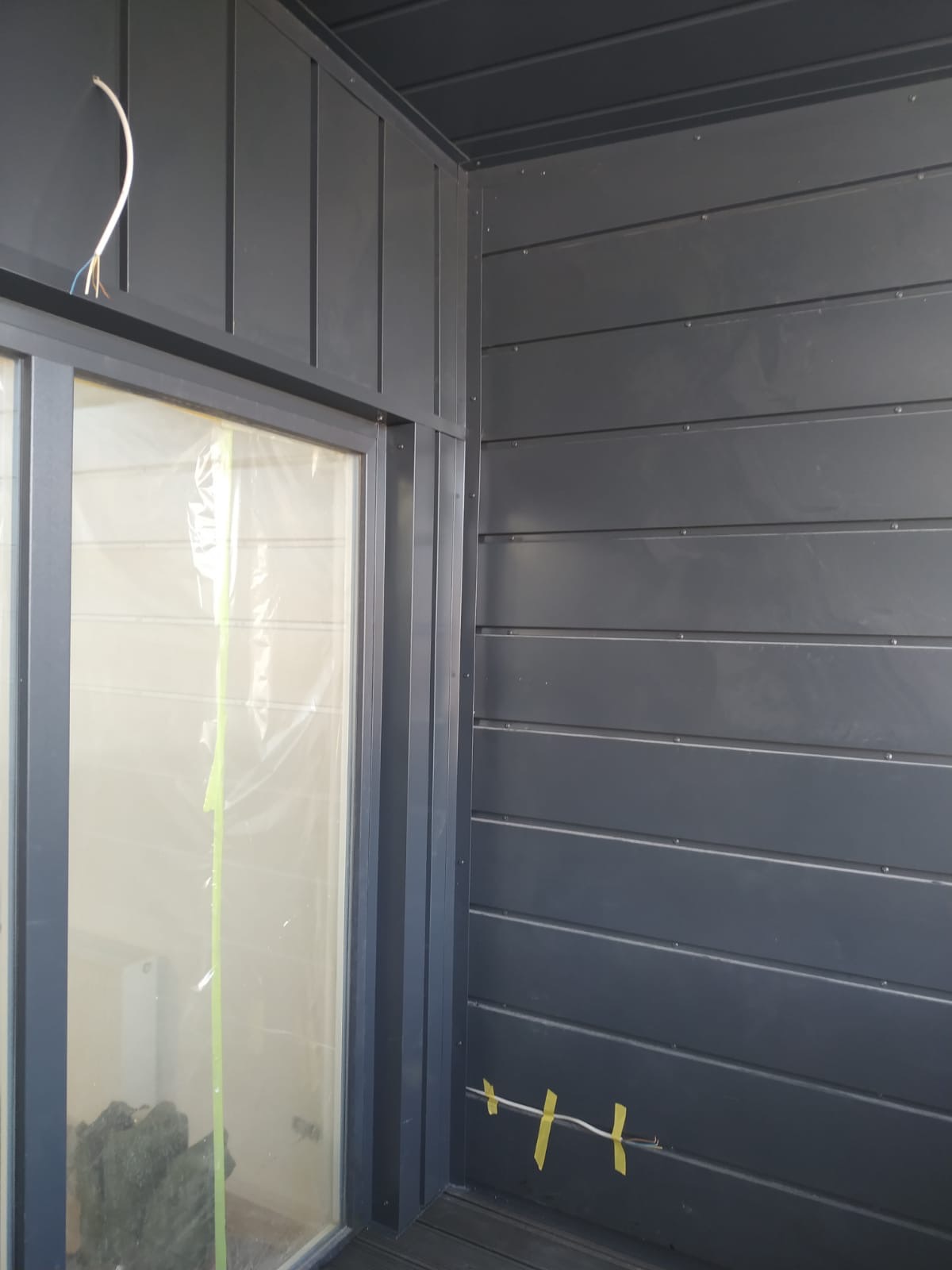 Szara elewacja z poziomych paneli, widoczne okno zabezpieczone folią i taśmą malarską, wystający kabel elektryczny przytwierdzony do ściany taśmą.