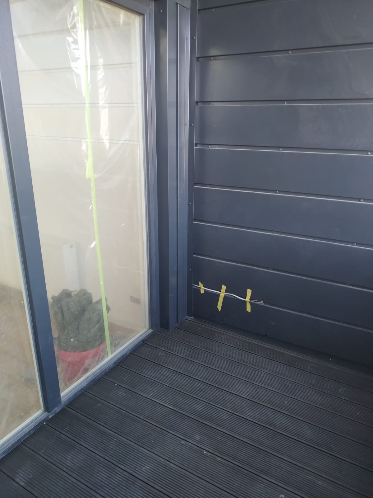 Fragment elewacji budynku z szarego sidingu, zabezpieczone folią okno, wystający kabel przymocowany taśmą malarską i podłoga z desek kompozytowych na balkonie