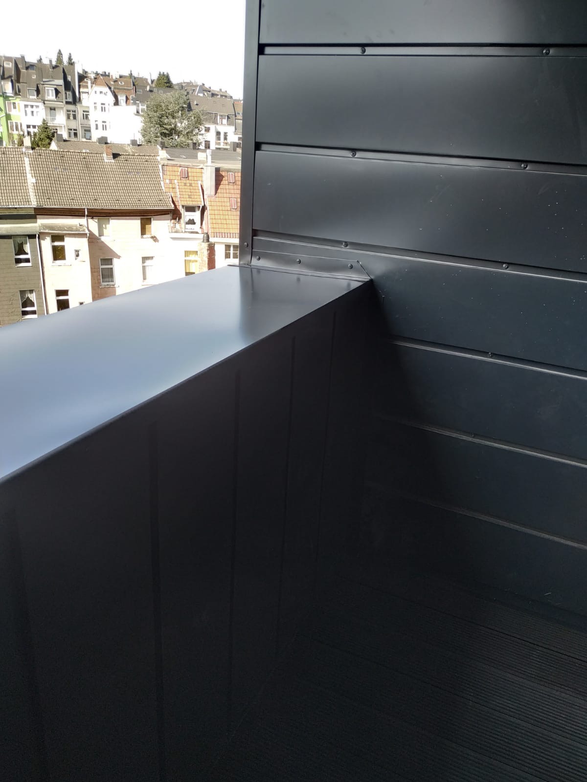 Szczegółowe ujęcie narożnika dachu z blachy w kolorze antracytowym, widoczne łączenia paneli i nity mocujące, w tle zabudowa miejska z dachami krytymi dachówką.