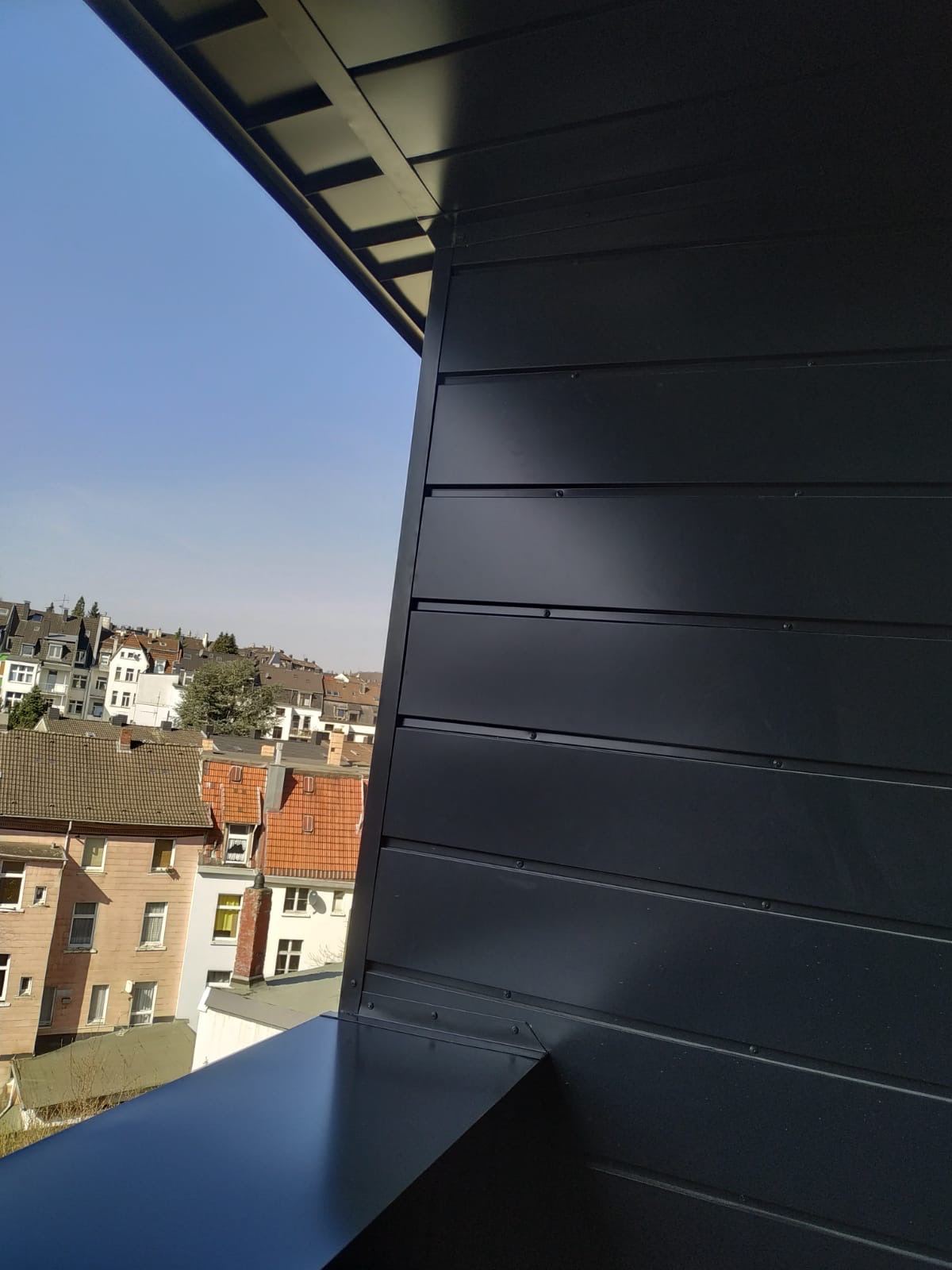 Fragment elewacji budynku z ciemnymi panelami, widok z góry na dachy sąsiednich budynków w słoneczny dzień.