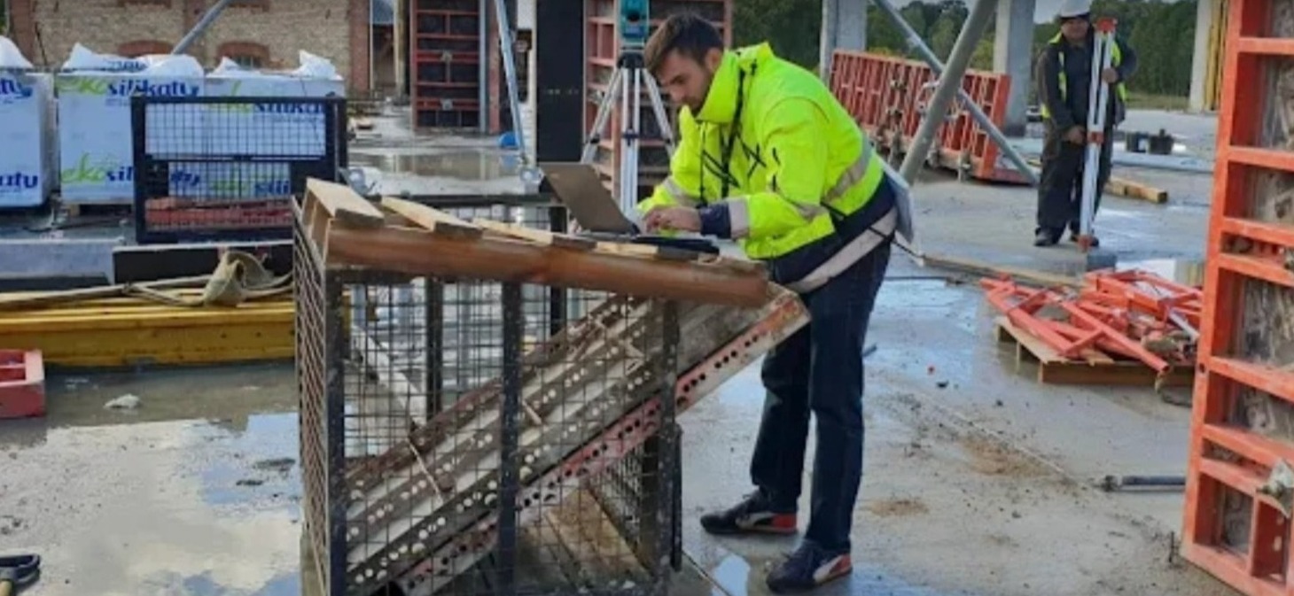 Pracownik w odblaskowej kurtce obsługuje laptopa na placu budowy, w tle teodolit i inny pracownik z łatą.