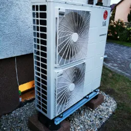 Biała pompa ciepła LG Therma V zamontowana na szarym cokole z kostki brukowej, otoczona ozdobnym żwirem, na tle elewacji budynku z widocznym fragmentem okna piwnicznego.