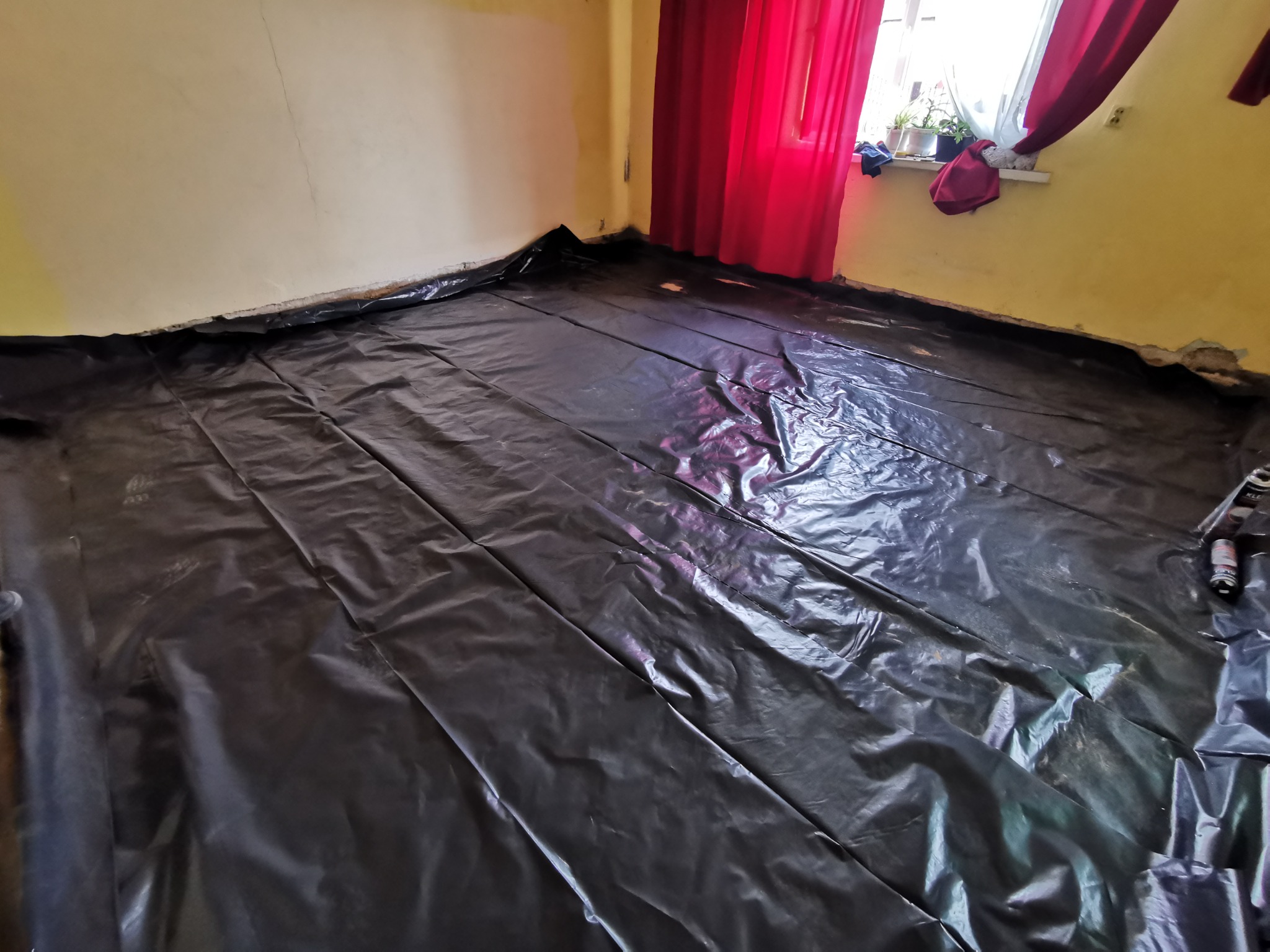 Przygotowanie do prac instalacyjnych: podłoga zabezpieczona czarną folią budowlaną w pomieszczeniu z żółtymi ścianami i czerwonymi zasłonami w oknie.