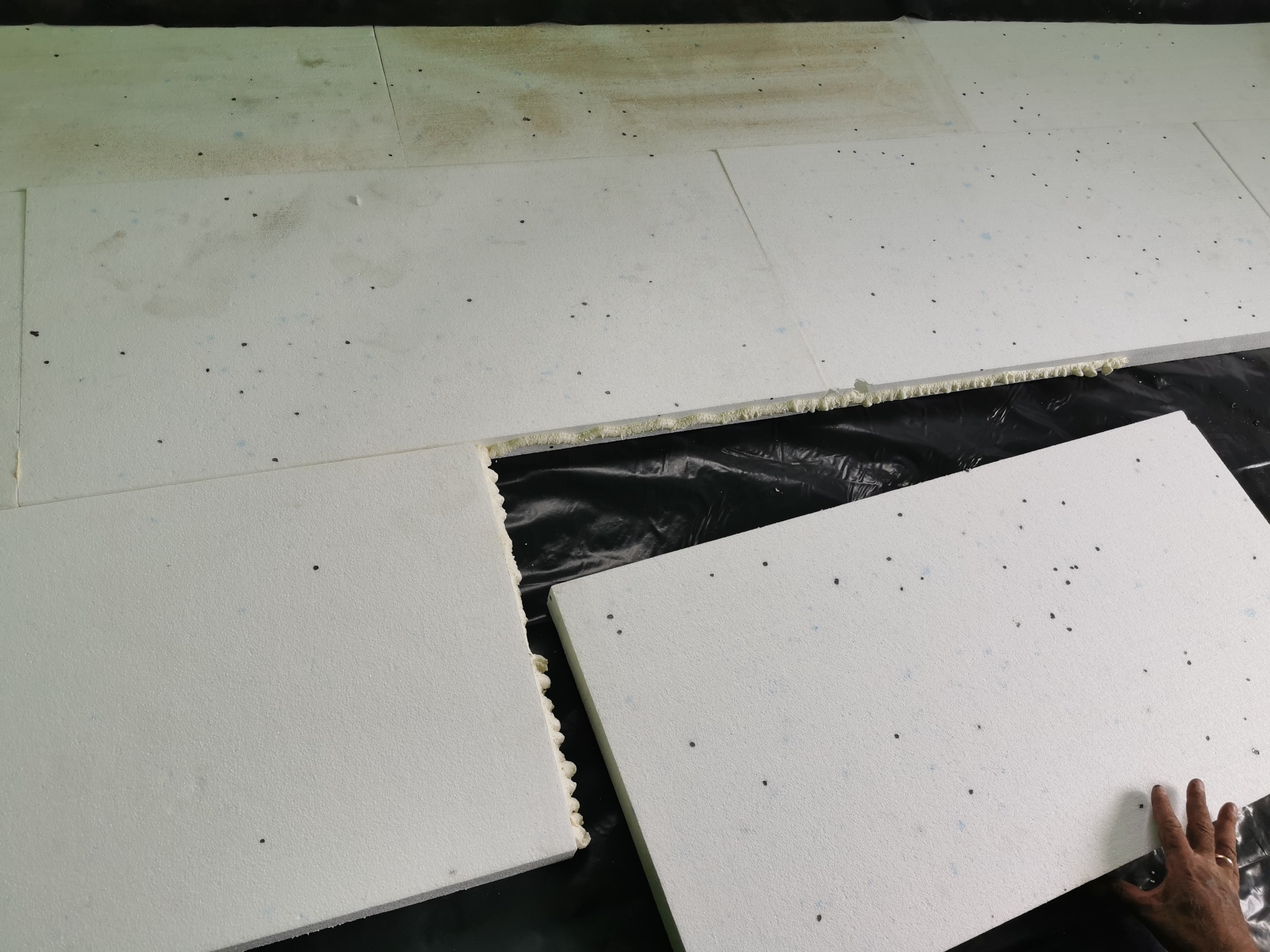 Układanie białych płyt izolacyjnych pod ogrzewanie podłogowe na czarnej folii, widoczna ręka z obrączką, szczeliny wypełnione pianką montażową.