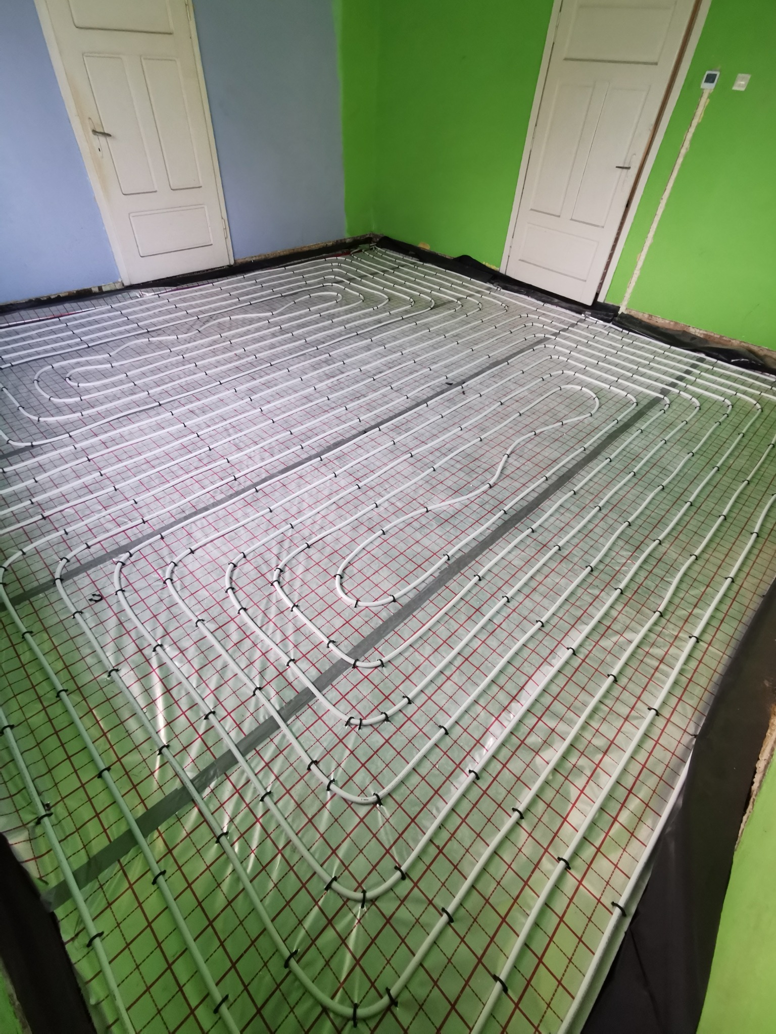 Białe rury ogrzewania podłogowego ułożone na folii z czerwoną siatką w pokoju z zieloną i niebieską ścianą, widoczne dwie białe drzwi.