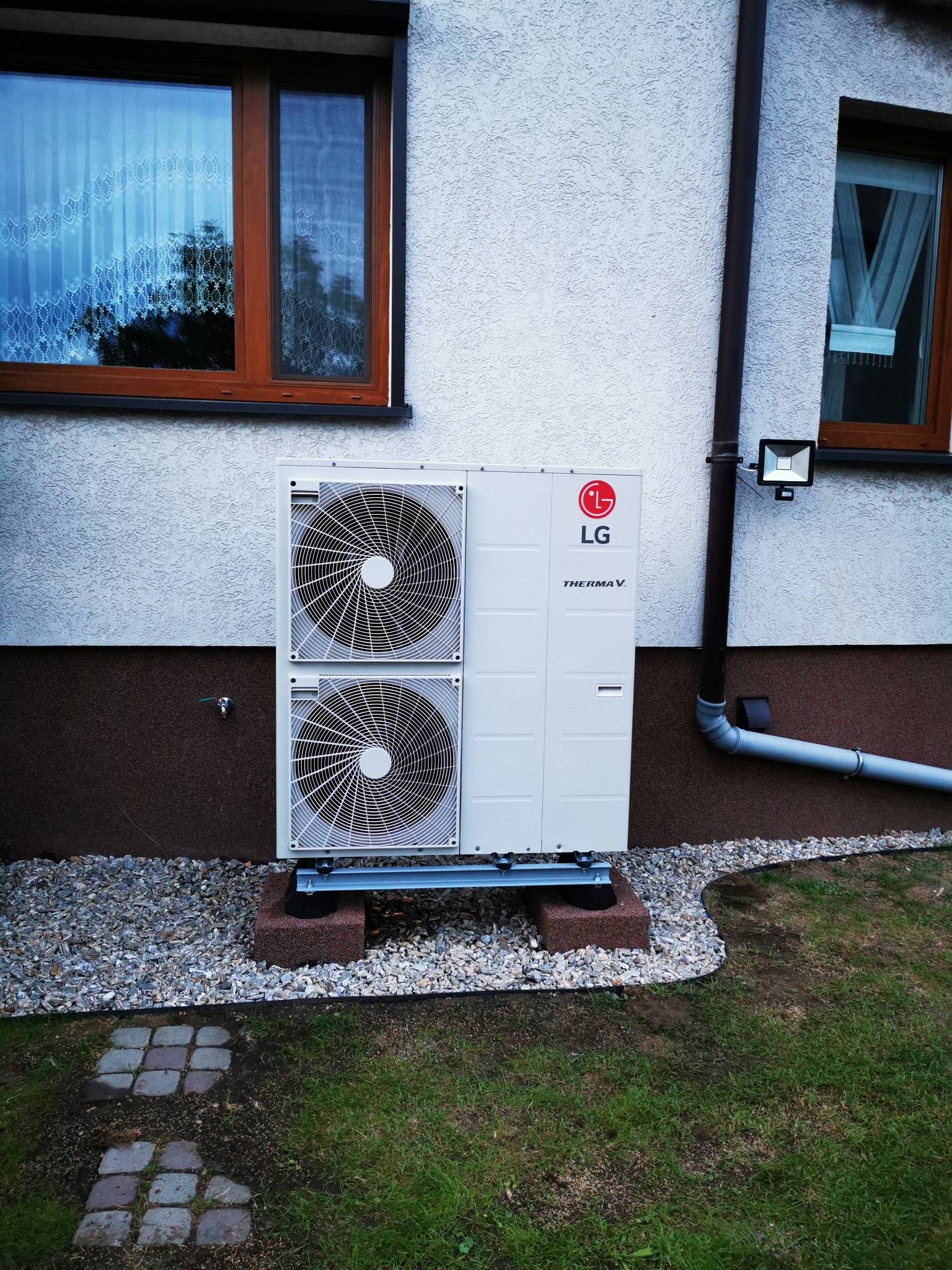 Biała jednostka zewnętrzna z dwoma wentylatorami, marki LG Therma V, zamontowana na podstawie z metalowych profili i betonowych bloczków, obok ściany domu z oknami i rynną.
