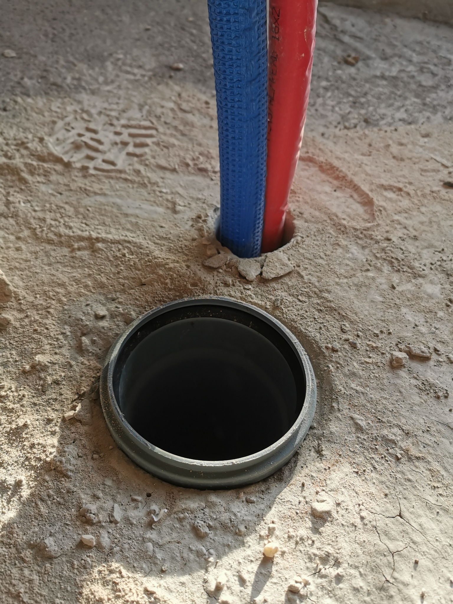 Przygotowanie do instalacji ogrzewania podłogowego: rury czerwone i niebieskie wystające z otworu w wylewce, obok czarna rura kanalizacyjna w ziemi, widoczne ślady butów na podłożu.