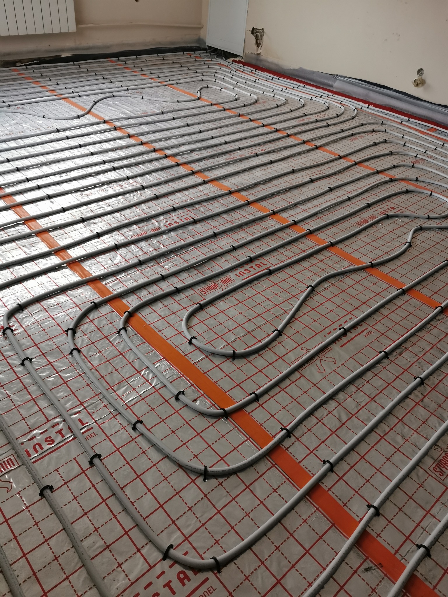 Równolegle ułożone rury ogrzewania podłogowego, przymocowane czarnymi uchwytami do folii z czerwoną siatką i pomarańczowymi paskami, widok z góry, fragment ściany w tle.