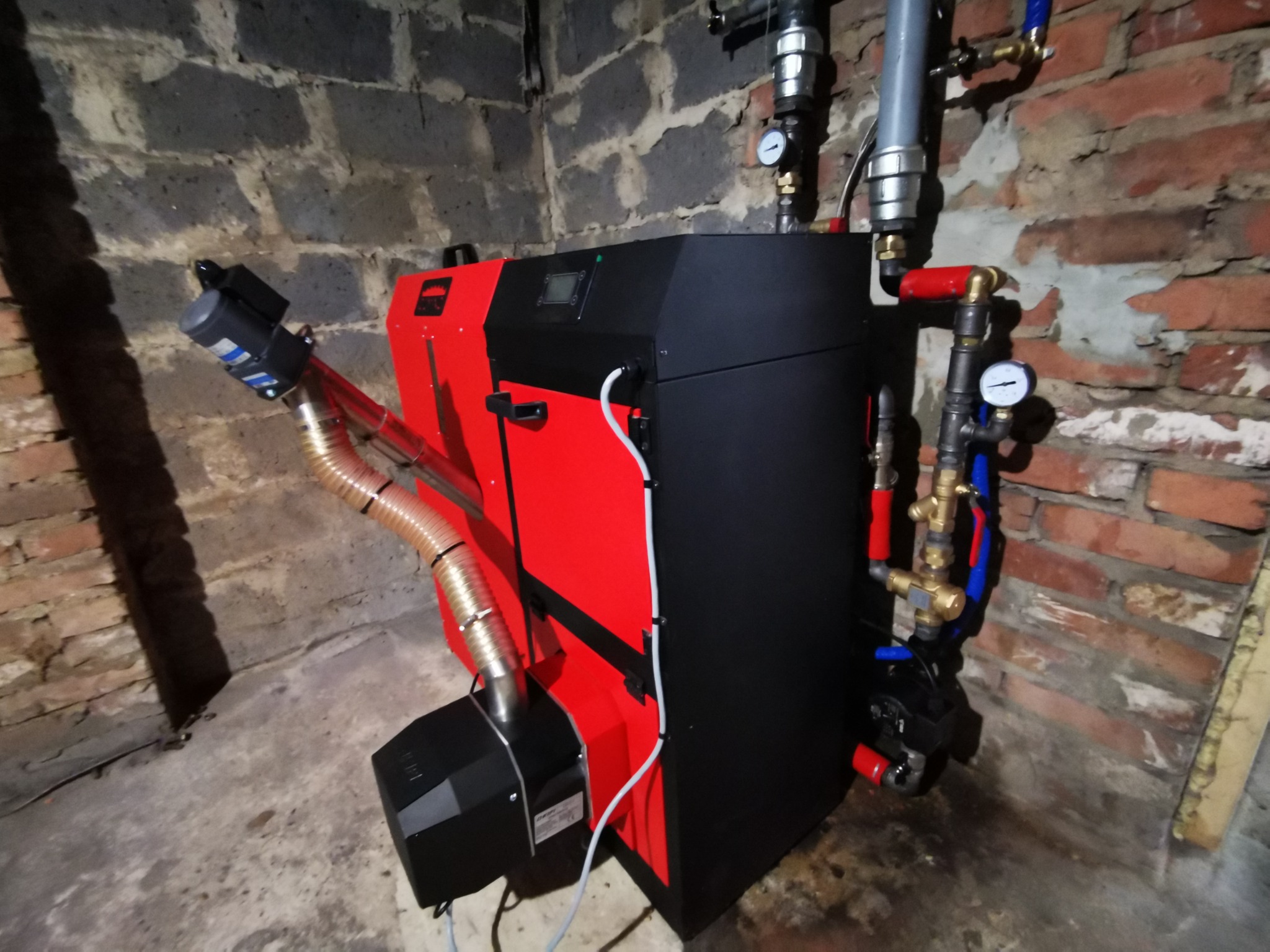 Czerwono-czarny piec CO z podajnikiem, zainstalowany w piwnicy z ceglanymi ścianami, widoczne rury i manometry.