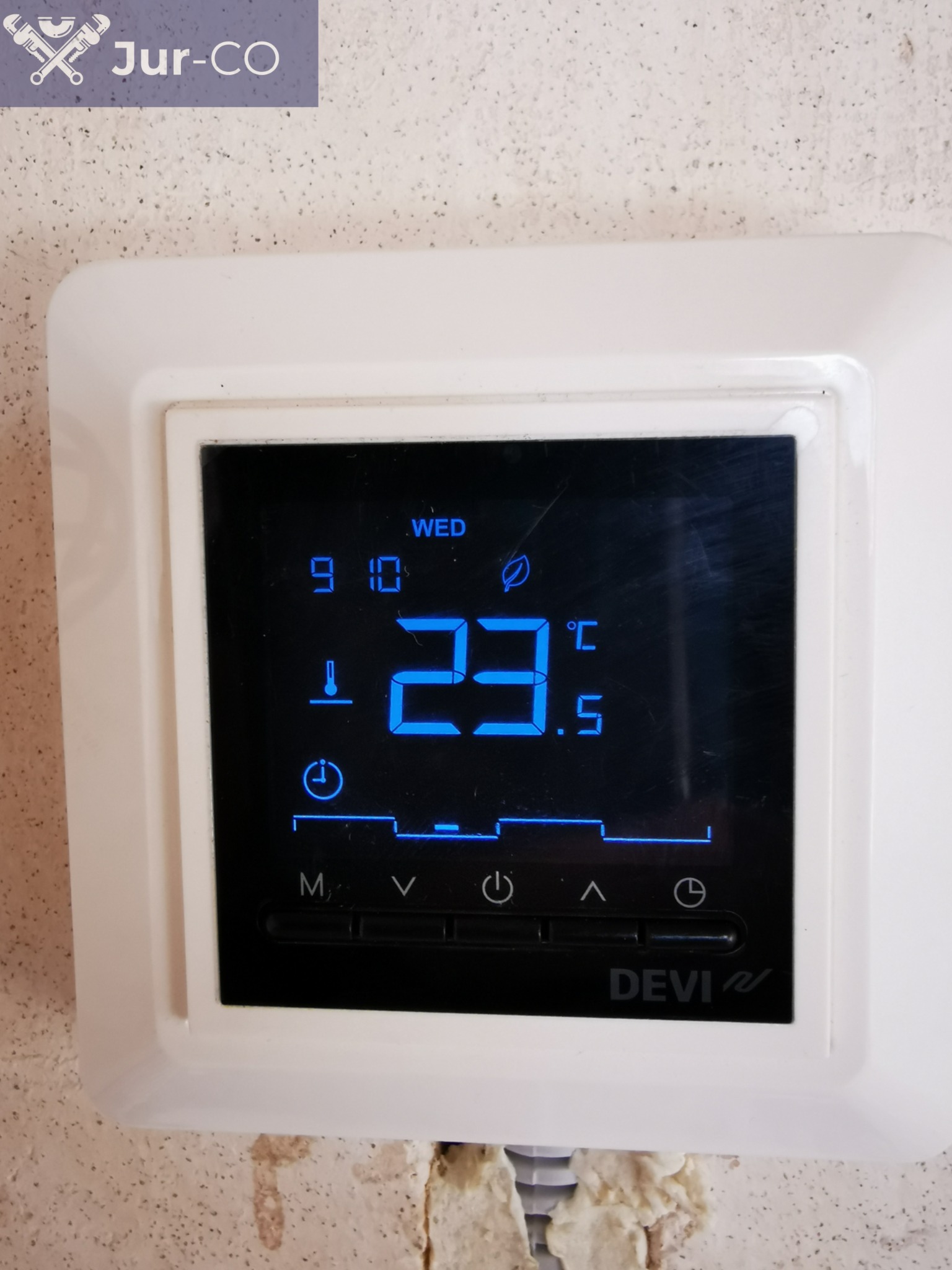 Biały termostat marki Devi zamontowany na ścianie, wyświetlający temperaturę 23.5 stopni Celsjusza, dzień tygodnia WED i godzinę 9:10.