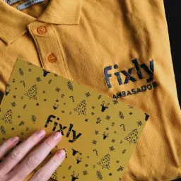 Żółta koszulka polo z haftowanym logo Fixly i napisem AMBASADOR, obok kartka z zimowymi motywami trzymana przez dłoń.