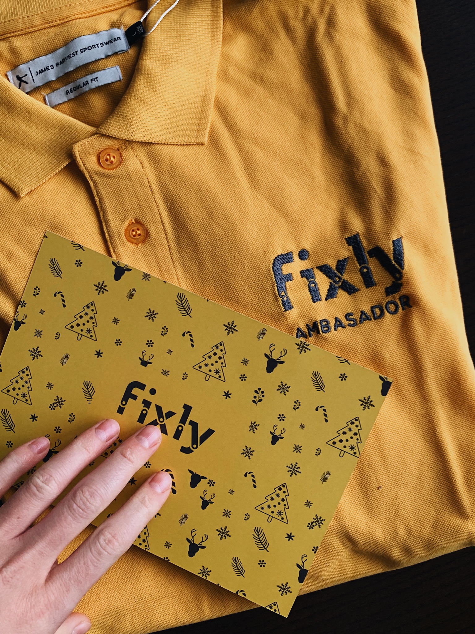 Żółta koszulka polo z haftowanym logo Fixly i napisem AMBASADOR, obok kartka z zimowymi motywami trzymana przez dłoń.
