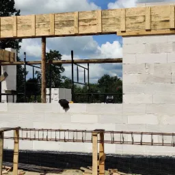 Wznoszenie ściany z betonu komórkowego z widocznym zbrojeniem i drewnianym szalunkiem nad otworem okiennym na tle zieleni.