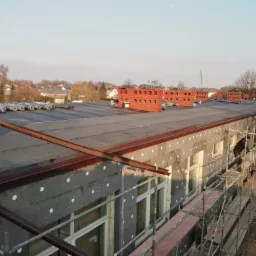 Budynek w trakcie ocieplania, widoczna szara izolacja ze styropianowymi kołkami, fragment dachu z rolkami papy, rusztowania i ceglane kominy na tle zabudowy jednorodzinnej.