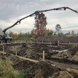 Wylewanie betonu na fundamenty domu jednorodzinnego za pomocą pompy do betonu z betoniarki, widoczni dwaj pracownicy w trakcie pracy.