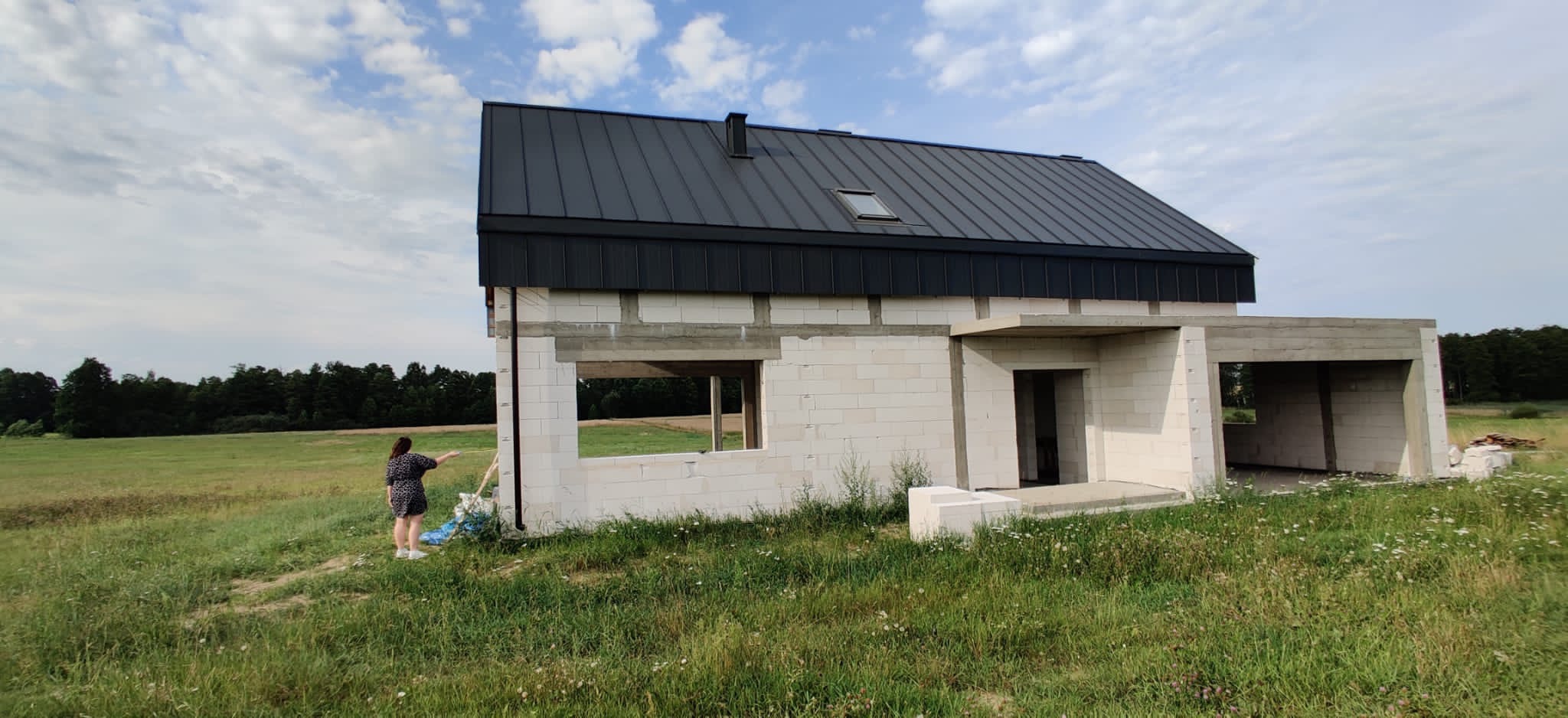 Niedokończony, parterowy dom z białych bloczków i czarnym dachem na tle łąki i lasu; kobieta w sukience stoi obok budynku, wskazując na niego.