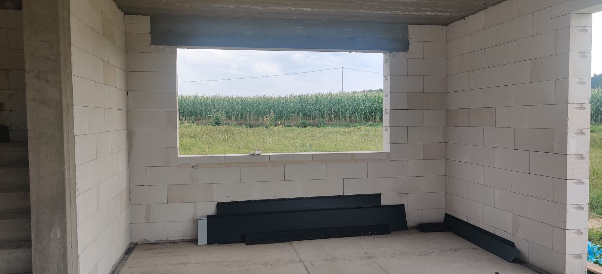 Wnętrze surowego domu z białych bloczków, widok przez otwór okienny na pole kukurydzy, czarne panele leżą na podłodze.