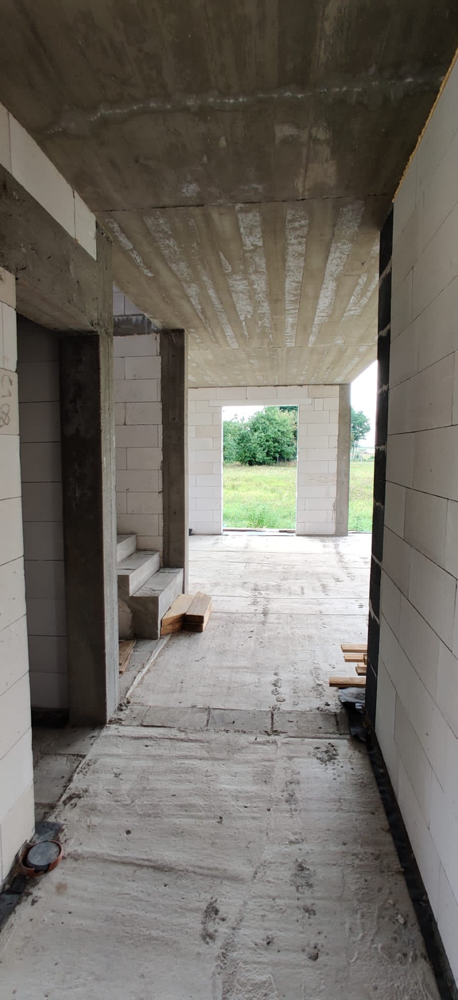 Wnętrze surowego domu w trakcie budowy, widoczne ściany z bloczków, betonowy strop z odciśniętymi deskami szalunkowymi, schody, oraz widok na zewnątrz przez otwór okienny.
