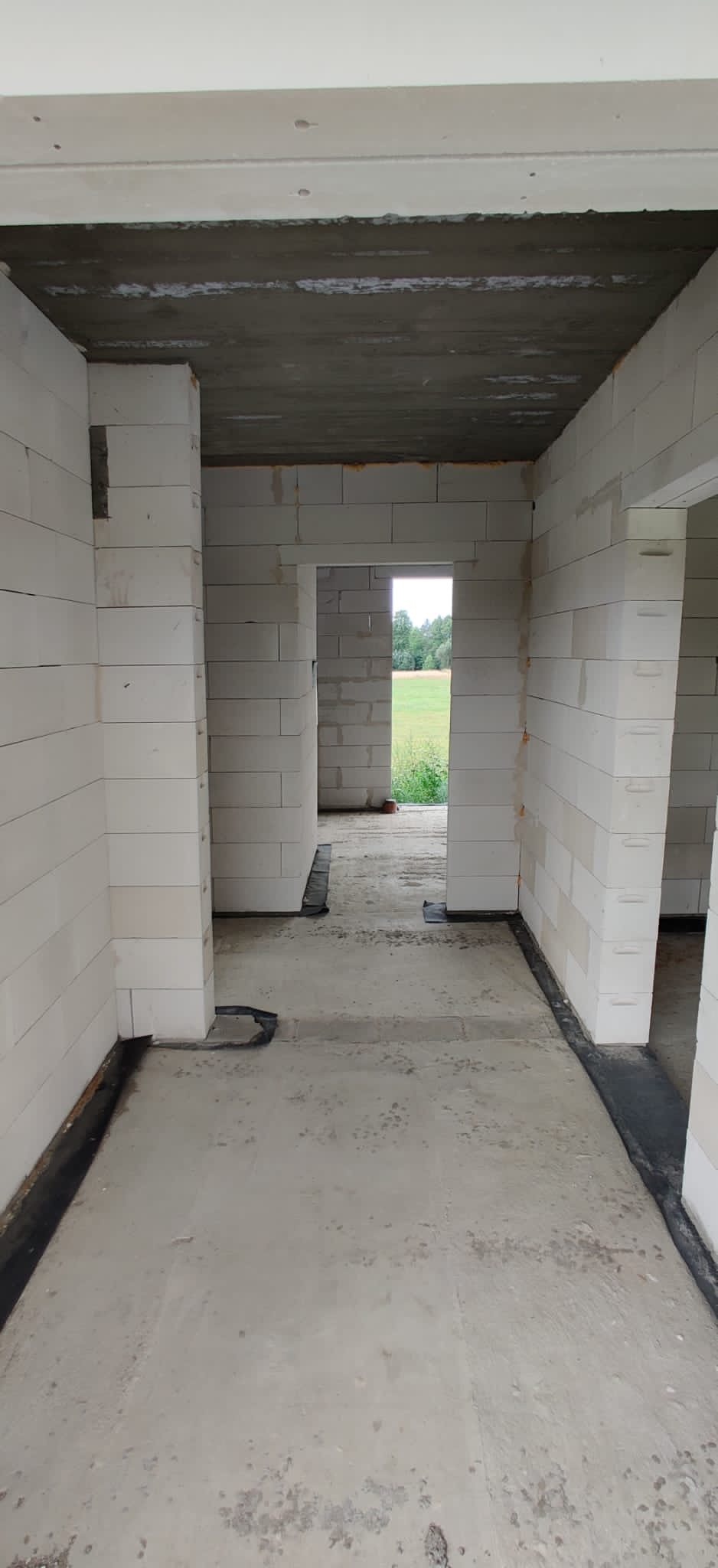 Wnętrze surowego budynku w trakcie budowy, ściany z białych bloczków, wylewka betonowa na podłodze, widoczne otwory drzwiowe i okienne z widokiem na zieleń.