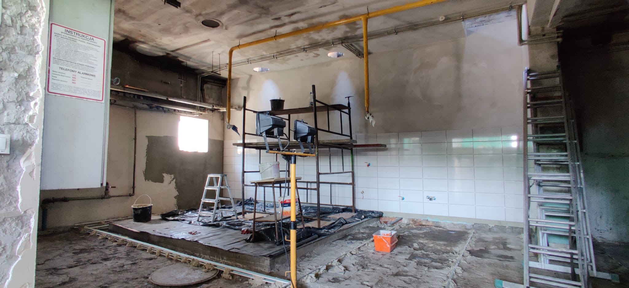 Wnętrze w trakcie remontu: rusztowanie, drabiny, odsłonięte podłoże, częściowo wykafelkowane ściany, żółte rury pod sufitem, tablica informacyjna na ścianie.
