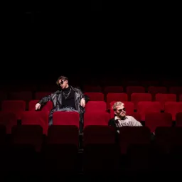 Dwóch mężczyzn w okularach przeciwsłonecznych siedzi na czerwonych fotelach w kinie lub teatrze, oświetlonych punktowo, sugerując sesję zdjęciową do teledysku.