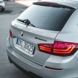 Tył srebrnego kombi BMW M550d z włączonymi światłami pozycyjnymi i widoczną tablicą rejestracyjną ZKL 30625 na tle szarej nawierzchni parkingu.