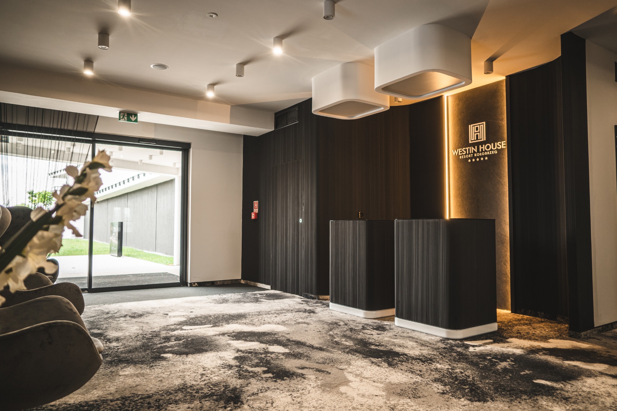 Nowoczesne lobby hotelowe z recepcją, oświetleniem LED i dywanem w abstrakcyjne wzory. Widoczny fragment wejścia i kwiatowa dekoracja.