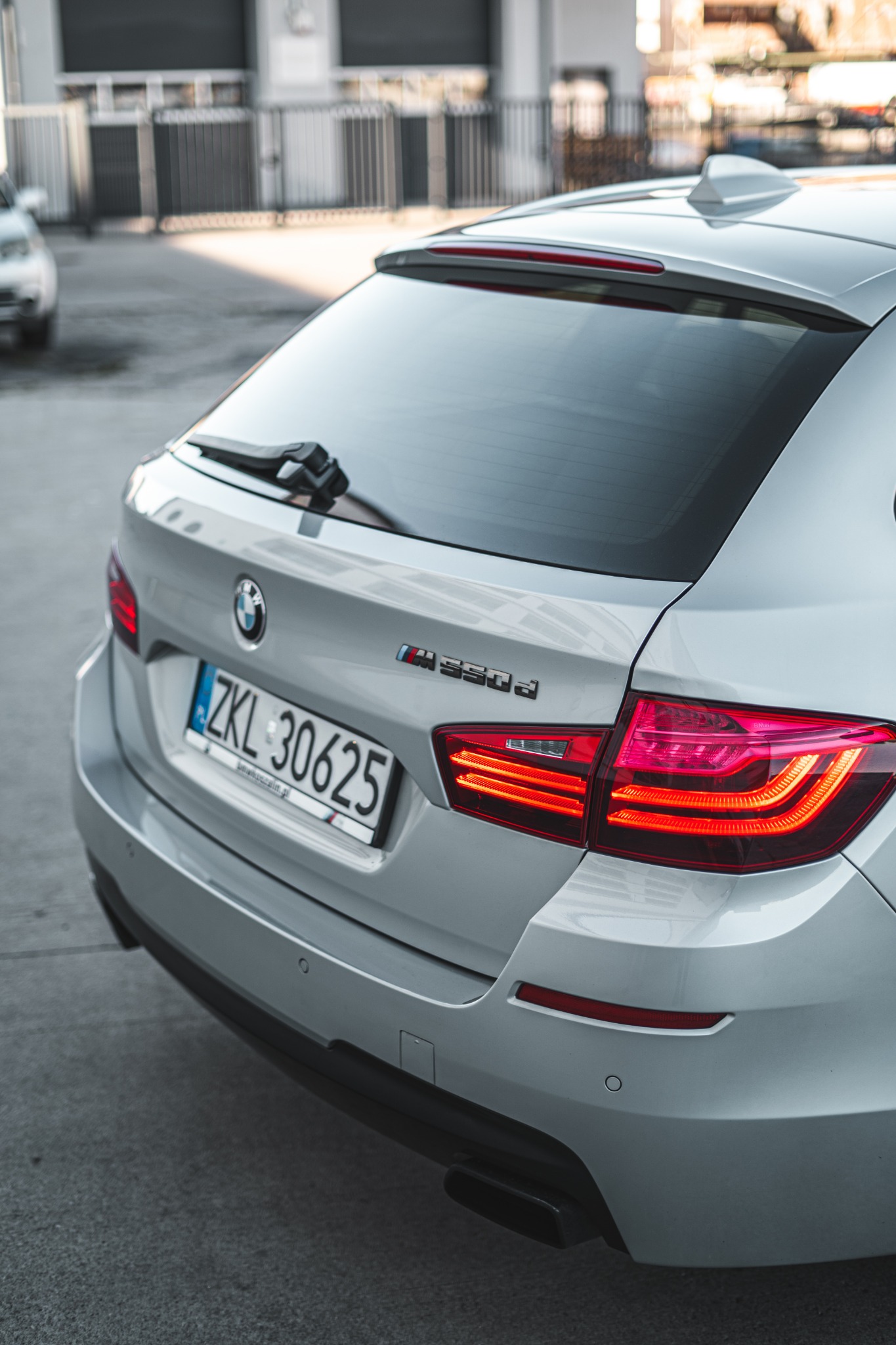 Tył srebrnego kombi BMW M550d z włączonymi światłami pozycyjnymi i widoczną tablicą rejestracyjną ZKL 30625 na tle szarej nawierzchni parkingu.