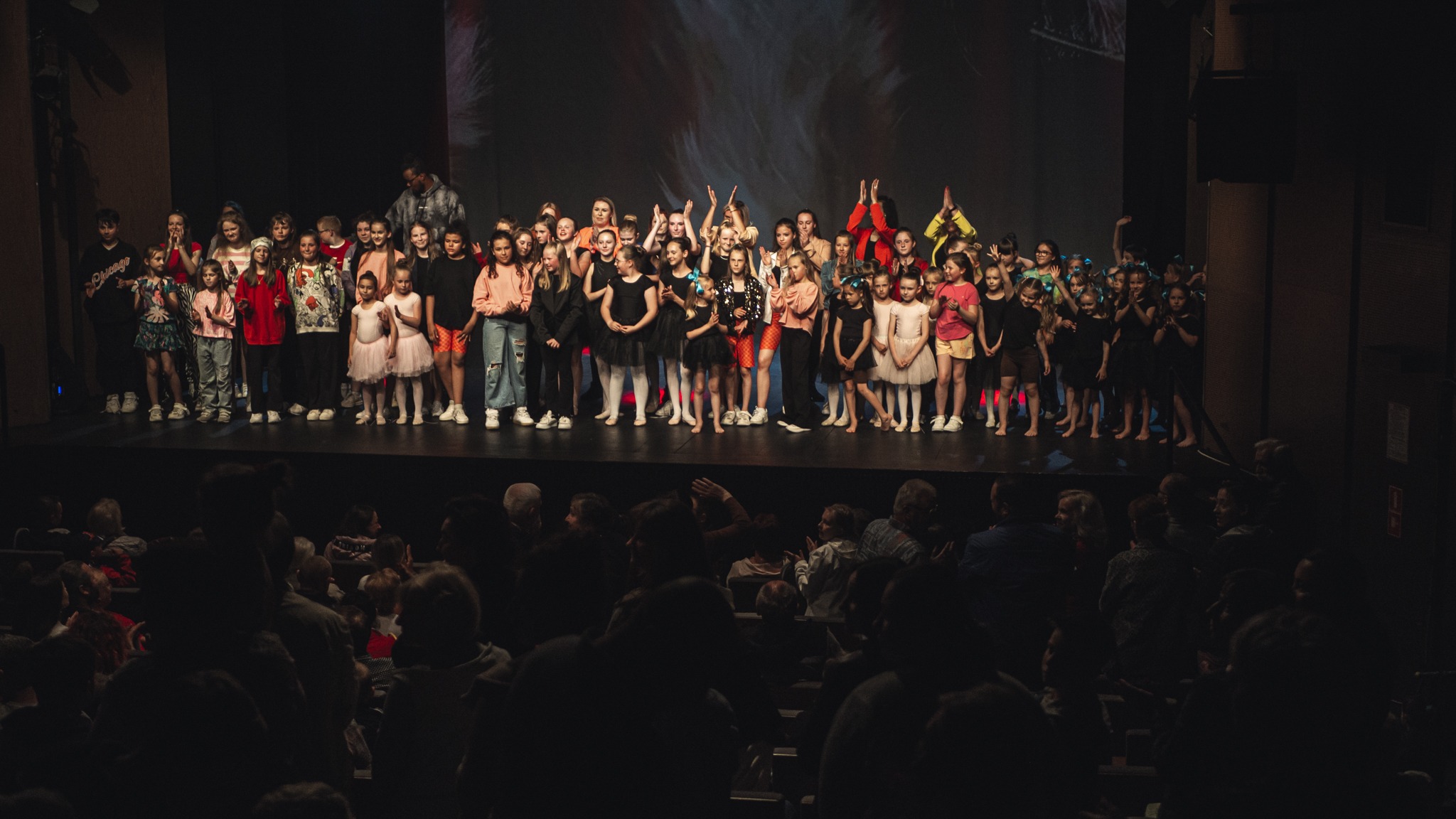 Duża grupa dzieci i młodzieży w różnorodnych strojach stoi na scenie, prawdopodobnie po występie tanecznym, publiczność siedzi na widowni.
