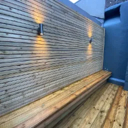 Nowoczesny balkon z drewnianą zabudową ścian i podłogi, oświetlony dwoma cylindrycznymi lampami ściennymi, z wbudowaną ławką, widok z dołu.