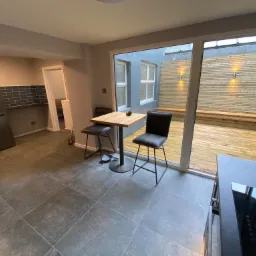 Nowoczesna kuchnia z szarymi płytkami podłogowymi, drewnianym stołem barowym i krzesłami, wychodząca na taras z drewnianą podłogą i oświetleniem ściennym.