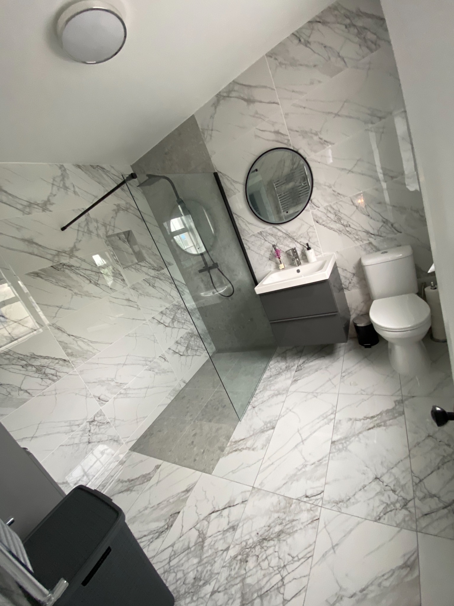 Nowoczesna łazienka z marmurowymi płytkami na ścianach i podłodze, szklaną kabiną prysznicową z czarnymi ramami, okrągłym lustrem nad szarą szafką umywalkową i wiszącą toaletą.