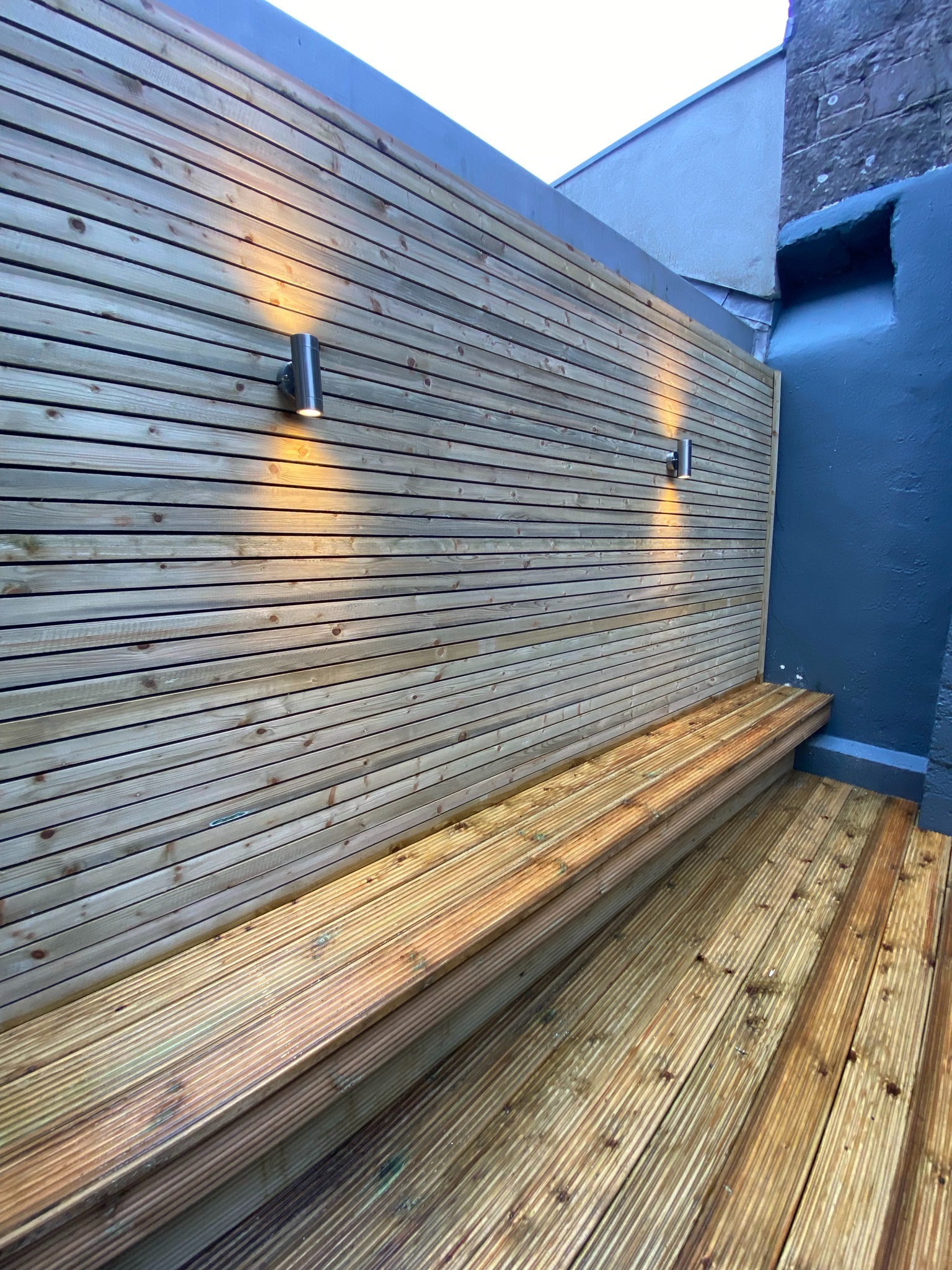 Nowoczesny balkon z drewnianą zabudową ścian i podłogi, oświetlony dwoma cylindrycznymi lampami ściennymi, z wbudowaną ławką, widok z dołu.