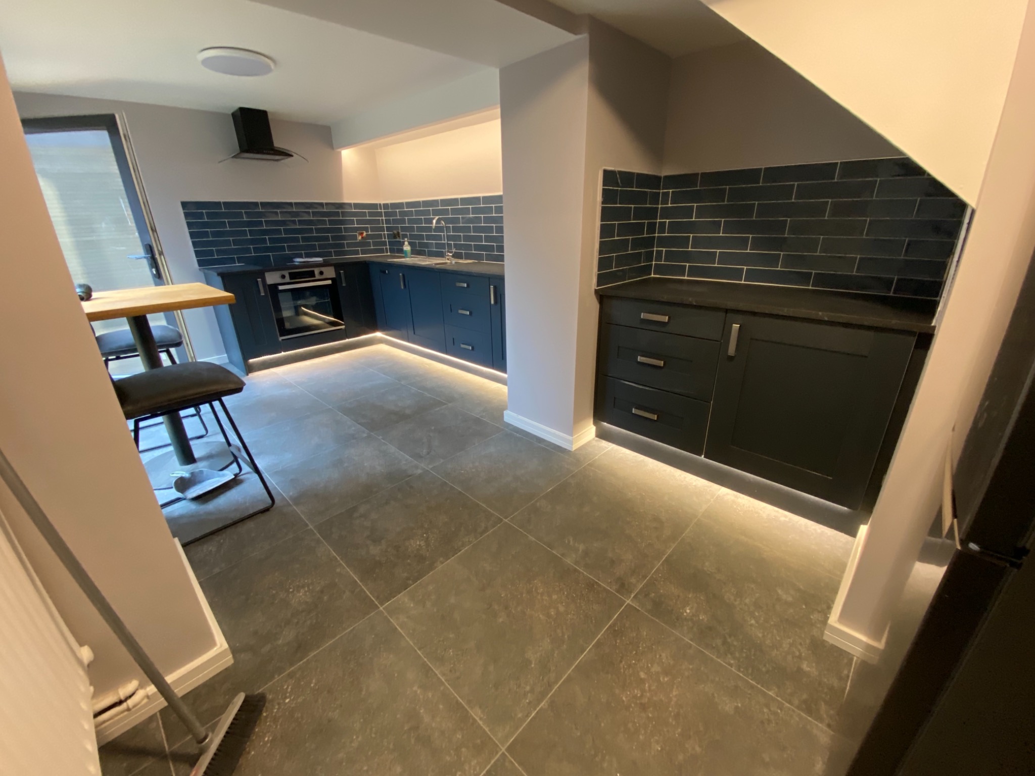 Nowoczesna kuchnia w odcieniach szarości i granatu z podświetleniem LED pod szafkami, widoczny stół barowy z krzesłami, ciemna podłoga z płytek imitujących kamień.