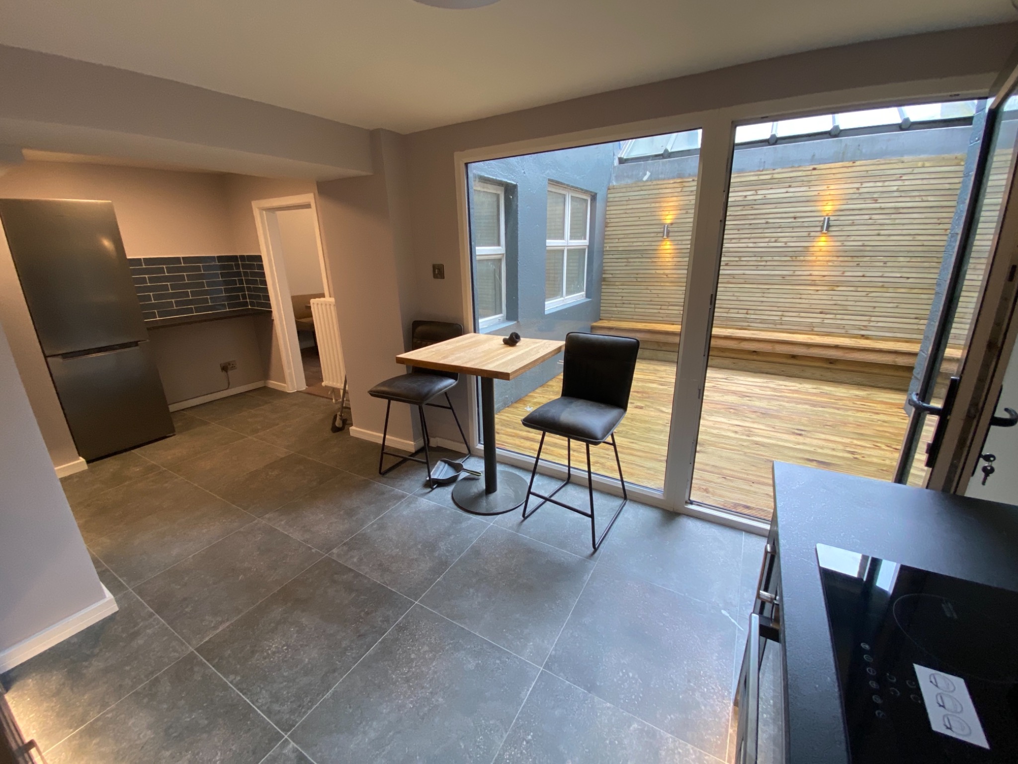 Nowoczesna kuchnia z szarymi płytkami podłogowymi, drewnianym stołem barowym i krzesłami, wychodząca na taras z drewnianą podłogą i oświetleniem ściennym.