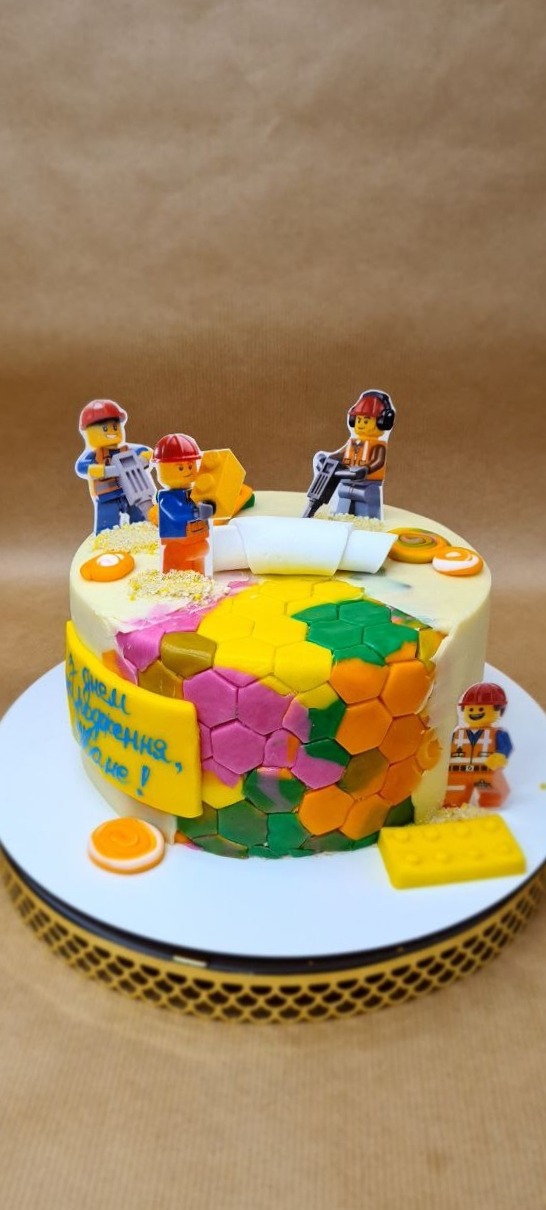 Okrągły tort urodzinowy z białym kremem, dekorowany kolorowymi, heksagonalnymi płytkami z masy cukrowej, figurkami Lego na górze i napisem w języku rosyjskim na żółtej tabliczce.