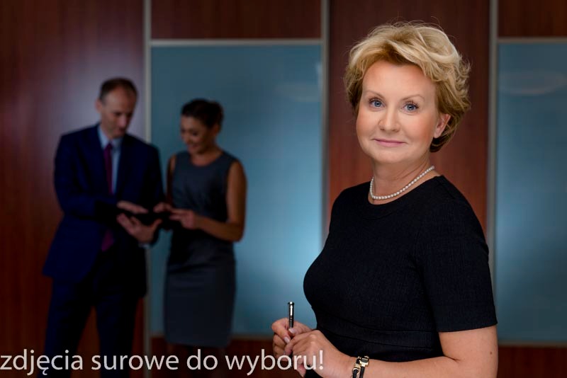 Portret kobiety w czarnej sukience, z perłami i zegarkiem, trzymającej długopis; w tle mężczyzna i kobieta przeglądający tablet w nowoczesnym biurze.