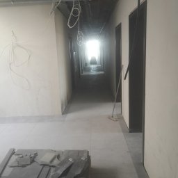 SeMoKo - Korytarz w trakcie remontu, widoczne kable pod sufitem, stos płytek na palecie i zamiatacz oparty o ścianę.