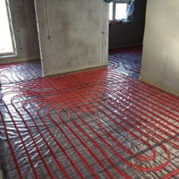 SeMoKo - Montaż ogrzewania podłogowego z czerwonych rur na folii izolacyjnej w surowym wnętrzu w Warszawie, widoczne szare ściany i okno.