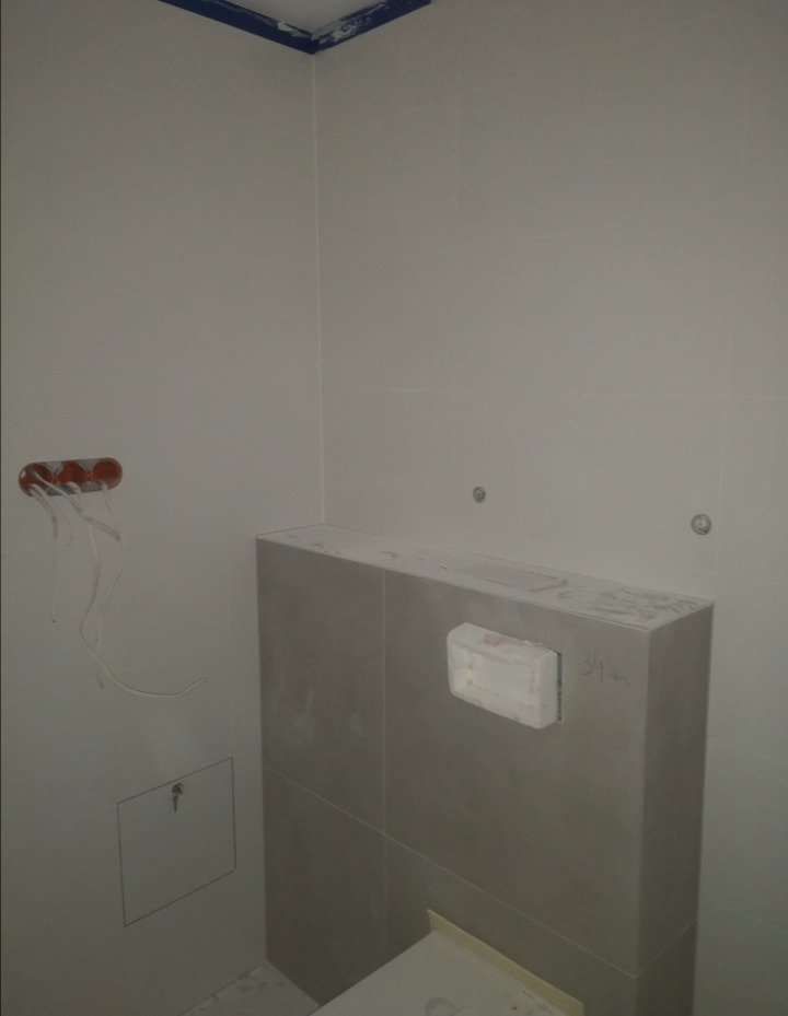 Szare płytki ścienne w trakcie montażu w łazience, widoczne przygotowane przyłącza elektryczne i fragment stelaża podtynkowego, ślady zaprawy na powierzchni płytek.