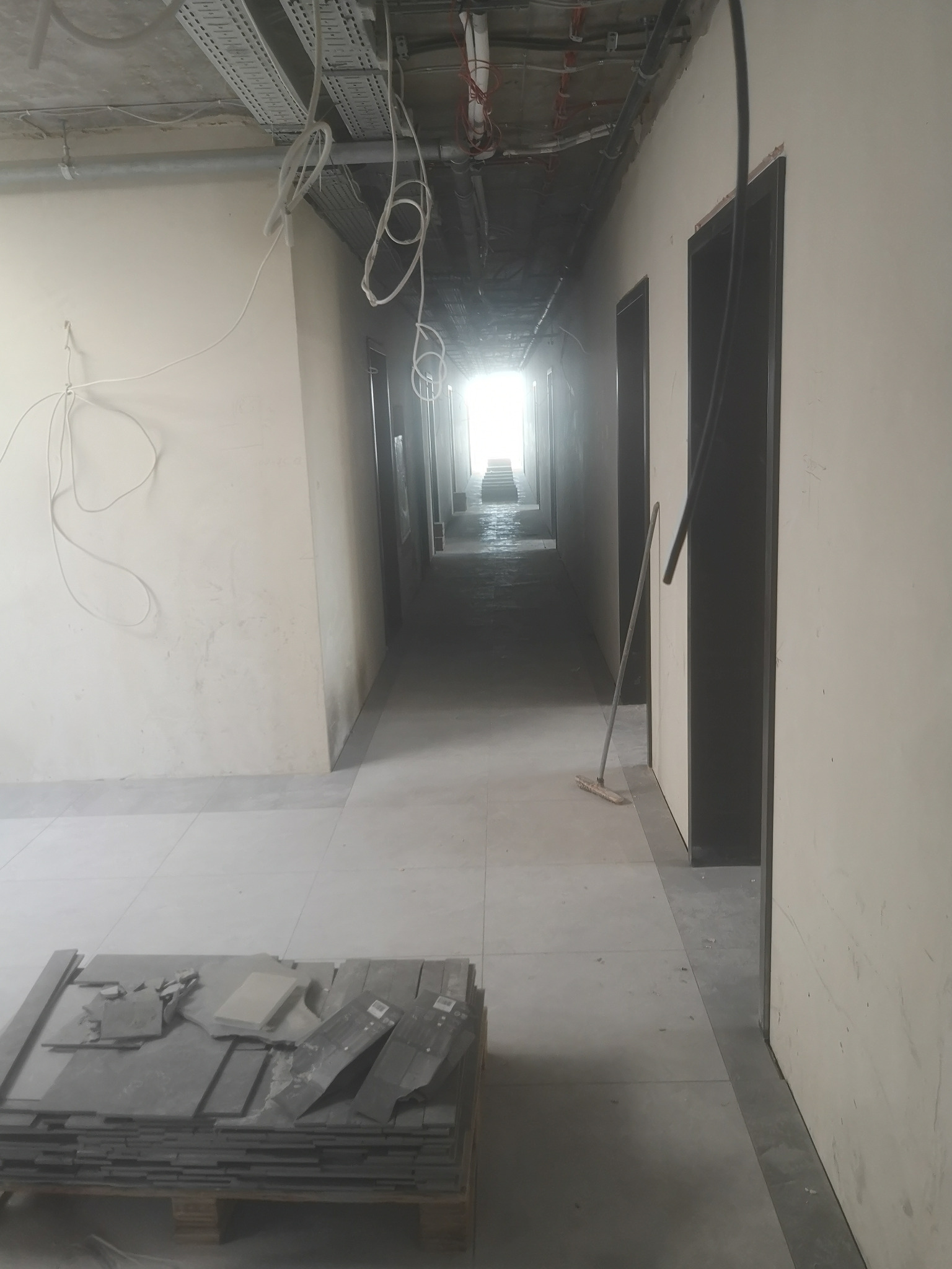 Korytarz w trakcie remontu, widoczne kable pod sufitem, stos płytek na palecie i zamiatacz oparty o ścianę.