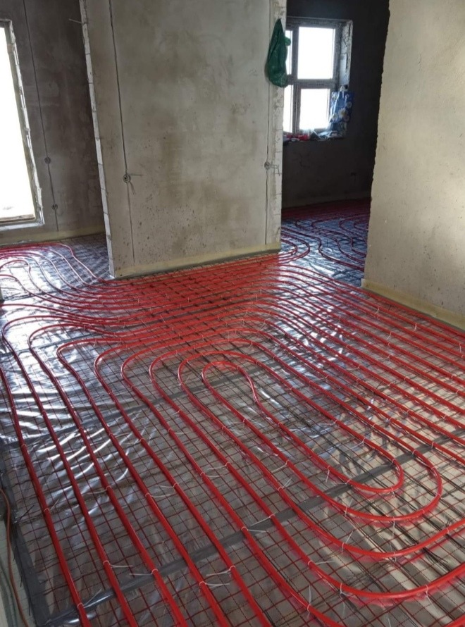 Montaż ogrzewania podłogowego z czerwonych rur na folii izolacyjnej w surowym wnętrzu w Warszawie, widoczne szare ściany i okno.