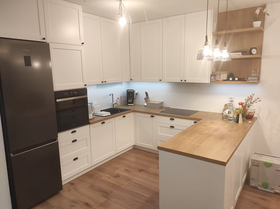 Jasna kuchnia z białymi szafkami, drewnianym blatem i czarnym sprzętem AGD, uzupełniona o drewnianą półkę z dekoracjami i wiszące lampy