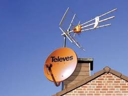 Pomarańczowa antena satelitarna marki Televes i antena kierunkowa VHF/UHF zamontowane na kominie z czerwonej cegły na tle błękitnego nieba.