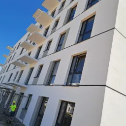 Nowoczesny budynek wielorodzinny z białymi ścianami i balkonami, widoczne rusztowania i pracownik w jaskrawej koszulce, błękitne niebo w tle.