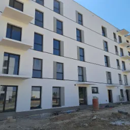 Nowoczesny, biały budynek wielorodzinny z czarnymi ramami okiennymi i małymi balkonami, widoczny w trakcie prac wykończeniowych na zewnątrz, z elementami rusztowania i pracownikami w tle.