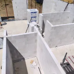 Wznoszenie ścian parteru domu z białych bloczków Solbet, widoczne drewniane szalunki stropu, teren budowy w słoneczny dzień.