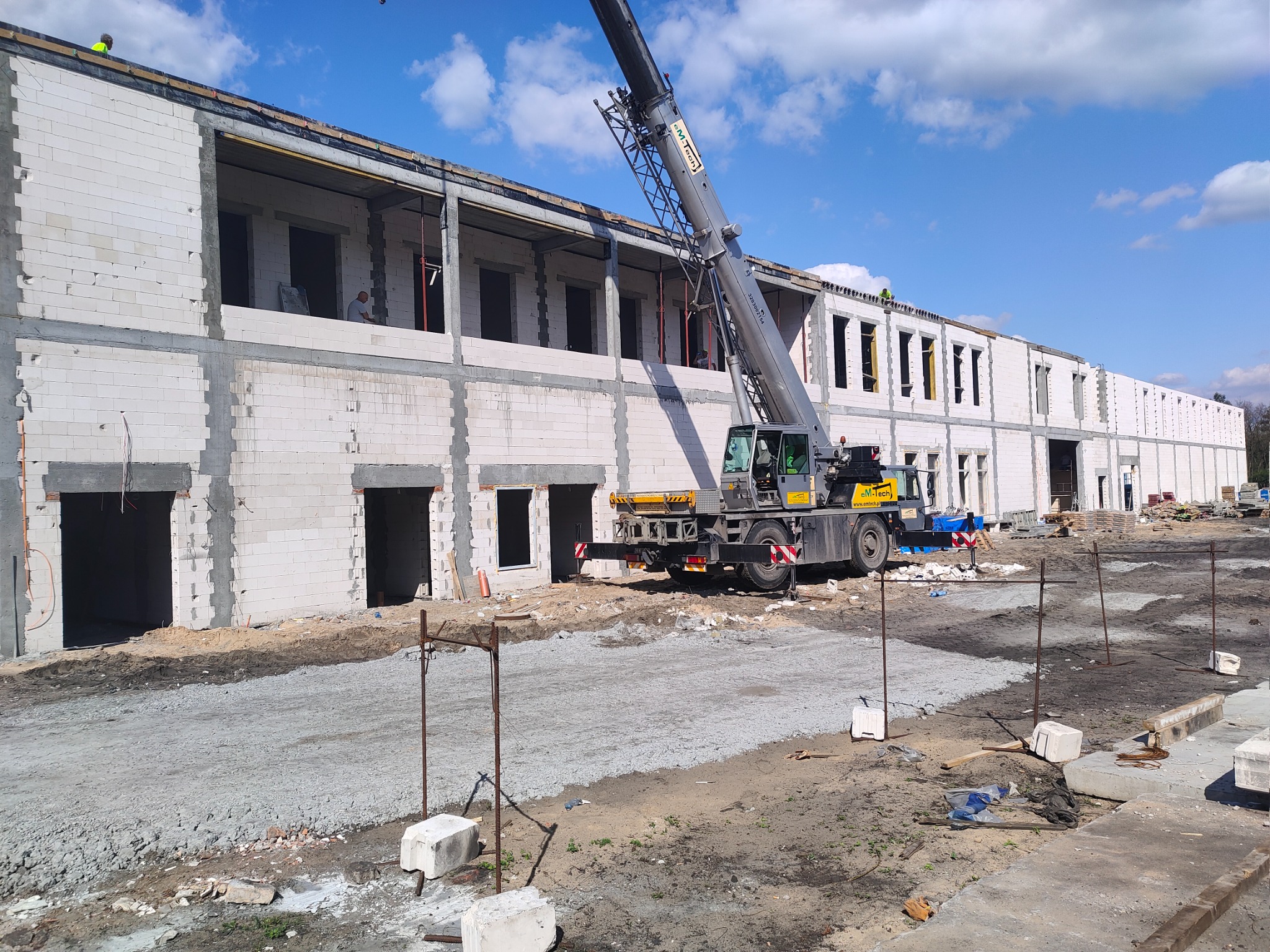 Wznoszenie dwukondygnacyjnego budynku z białych bloczków, widoczny dźwig budowlany i teren przygotowany pod wylewkę, błękitne niebo z chmurami w tle.