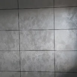 Ściana pokryta dekoracyjnym szarym tynkiem strukturalnym podzielona czarnymi listwami na regularne kwadraty, widoczny fragment okna z drewnianym parapetem.